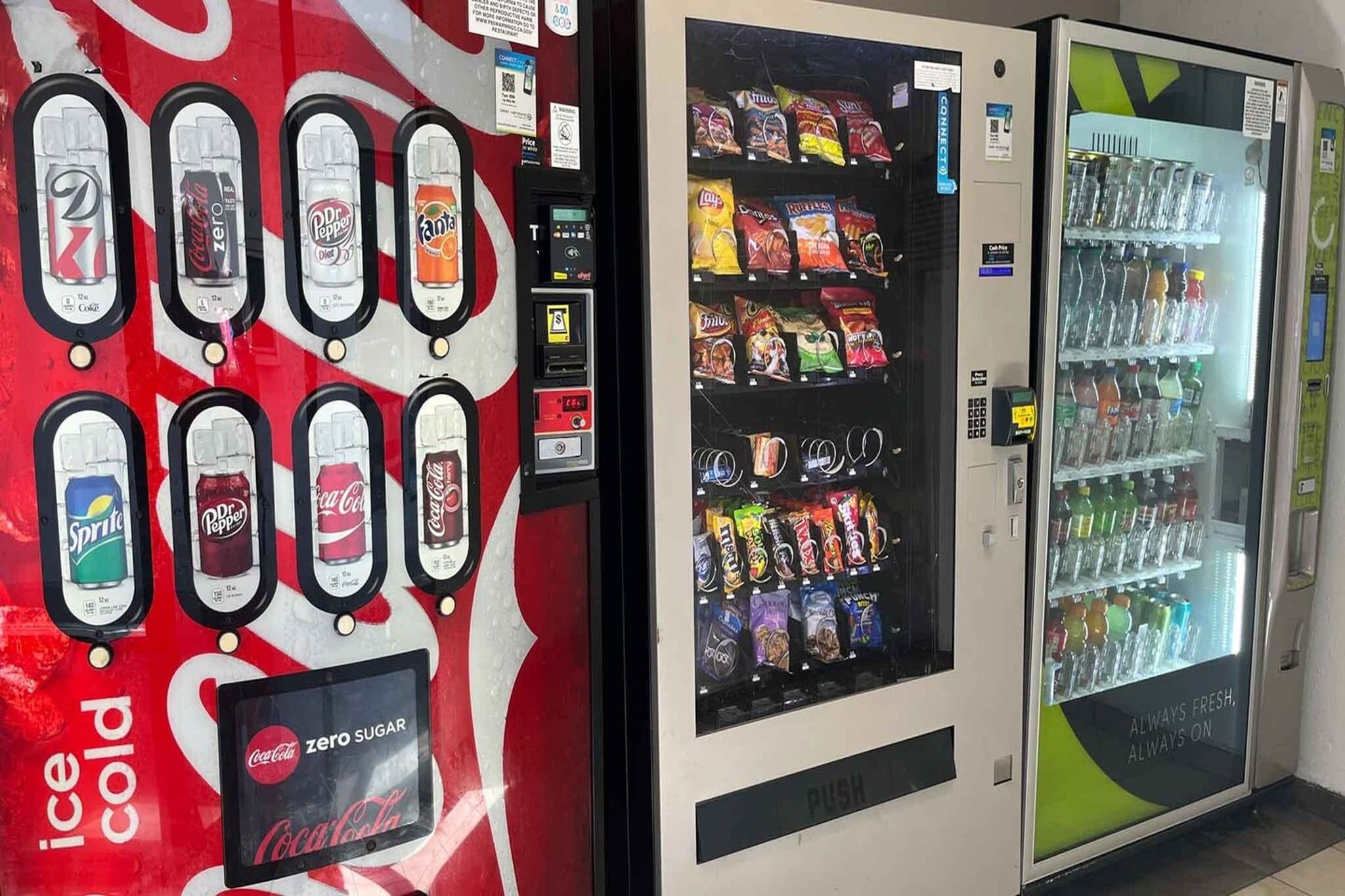 A vending machine with various beverage options, a Coca-Cola branded machine, and a refrigerator stocked with snacks and drinks.