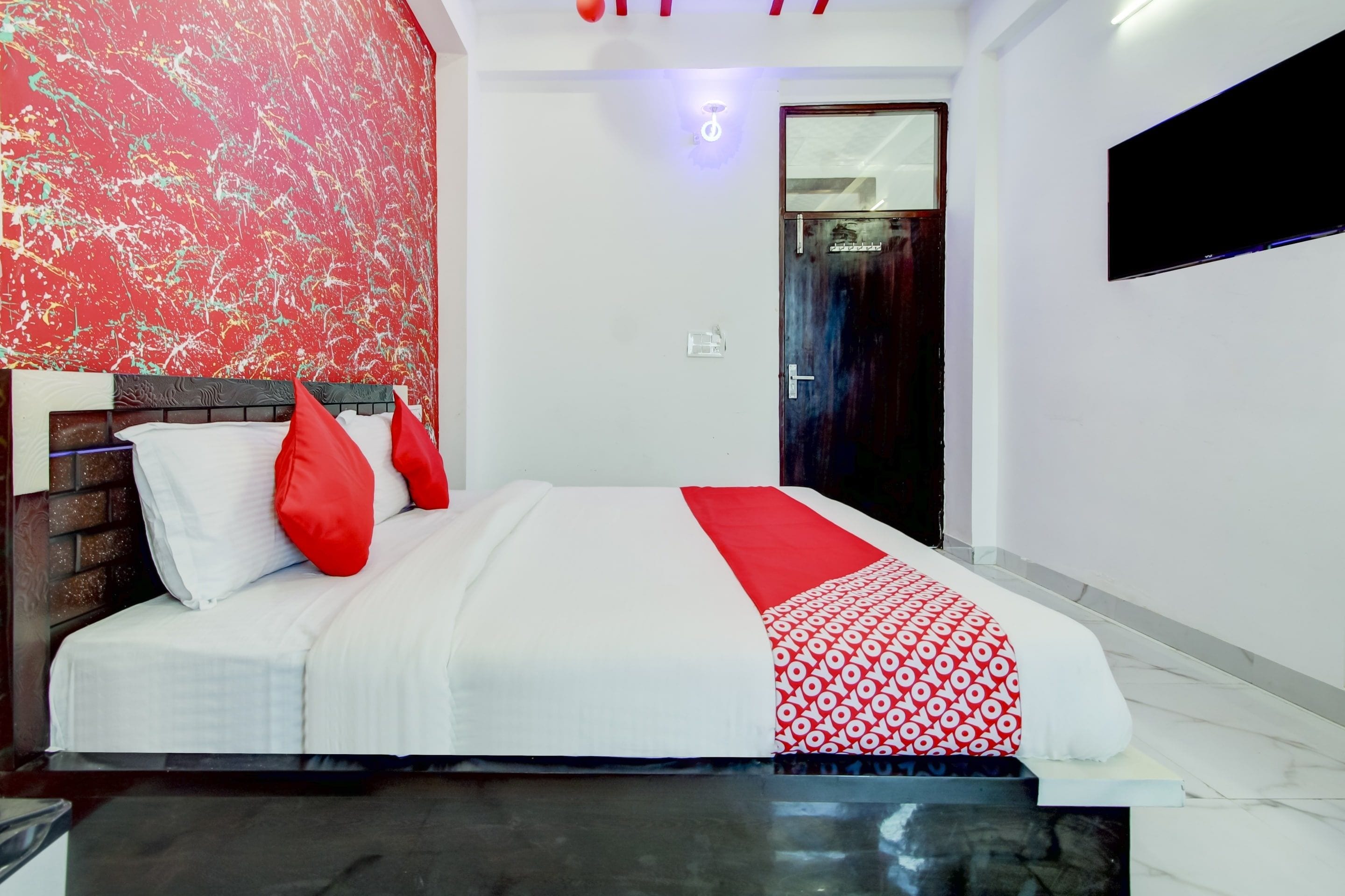 Une chambre d’hôtel avec un lit, une couvre-lit à motifs rouges et blancs, un oreiller rouge, une tête de lit à motifs de briques, un mur blanc et une porte avec un miroir.