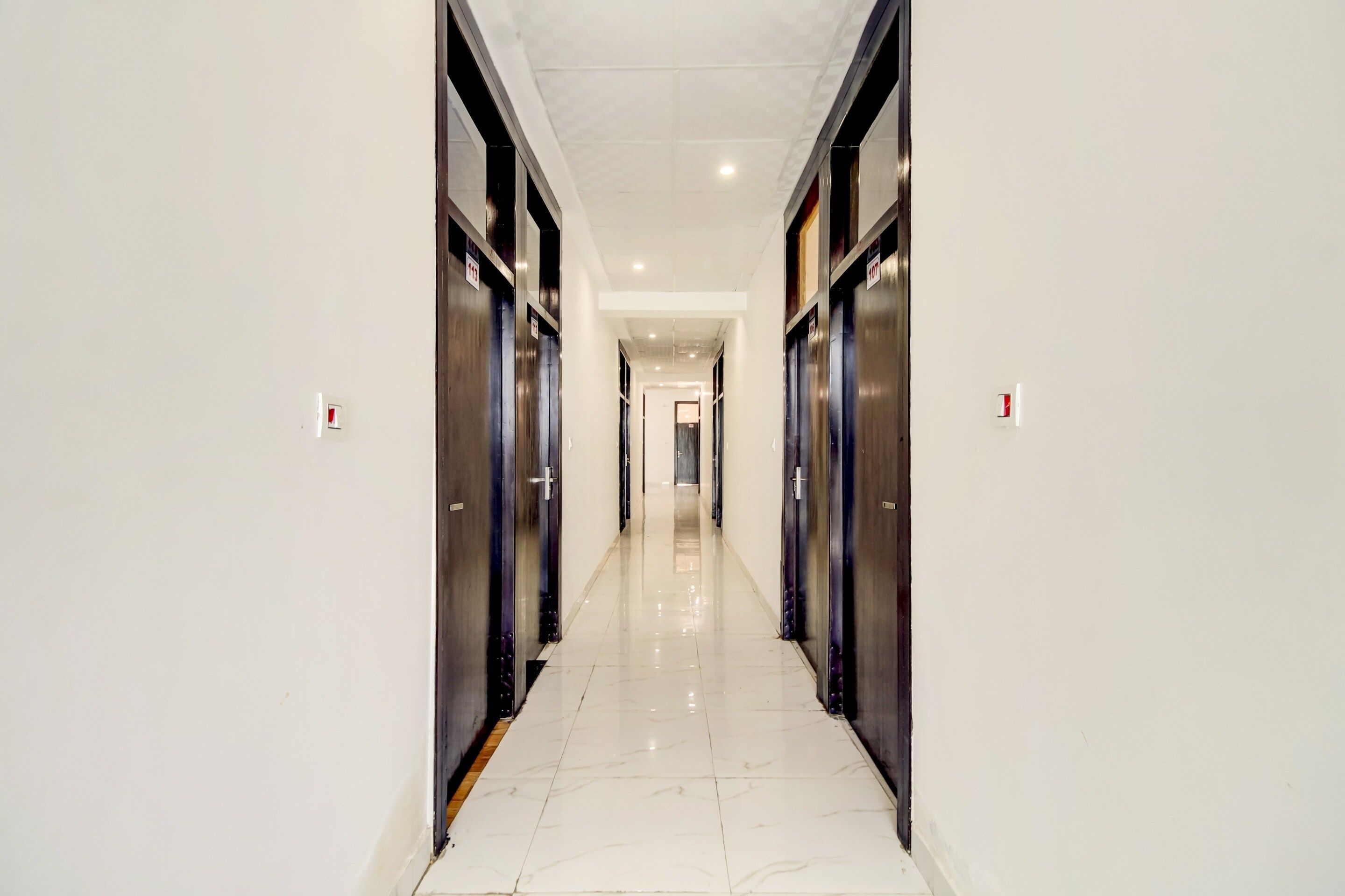 A corridor with a shiny tiled floor, flanked by dark-colored doors, leading to a bright room at the end.