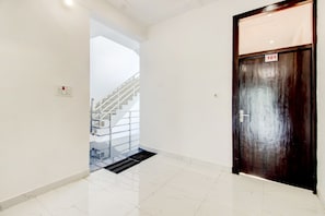 A hallway with a white tiled floor, a wooden door with a number plate, and a staircase with a glass railing.