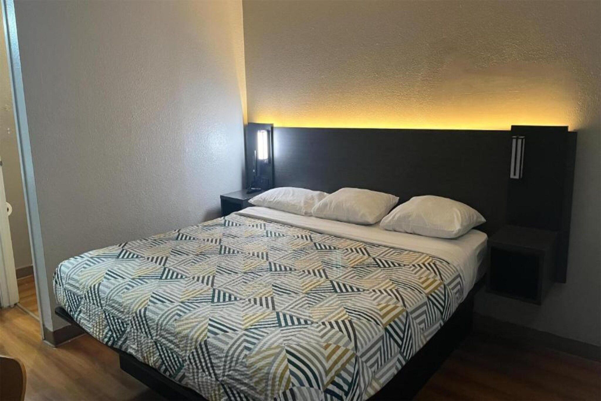 Un llit amb un estovat estampat, una taula de nit amb una làmpada i una prestatgeria penjada a la paret.
