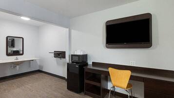 A hotel room with a flat-screen TV mounted on the wall, a small desk with a chair, a sink, and a mirror.