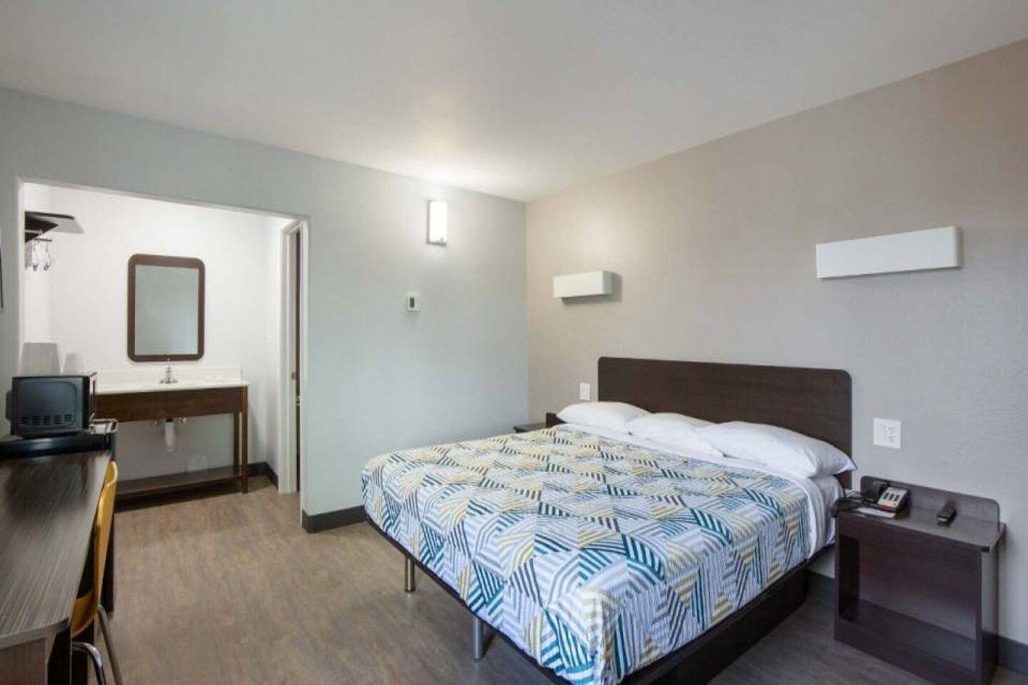 A hotel room with a bed, a desk, a chair, a mirror, and a bedside table with a phone.