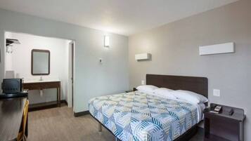 A hotel room with a bed, a desk, a chair, a mirror, and a bedside table with a phone.