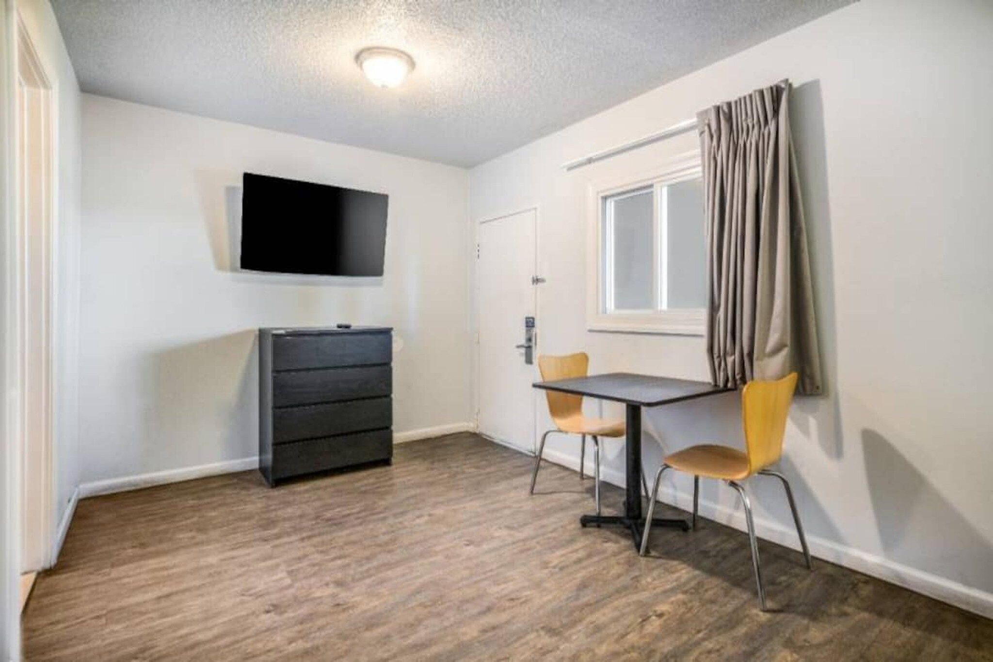 Een kamer met een wandgemonteerde flatscreen televisie, een klein zwart dressoir, een tafel met twee stoelen en een raam met gordijnen.
