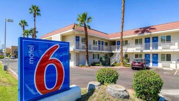 Une enseigne de motel avec le chiffre 6, un bâtiment à deux étages doté de balcons, de palmiers et d’une voiture garée.