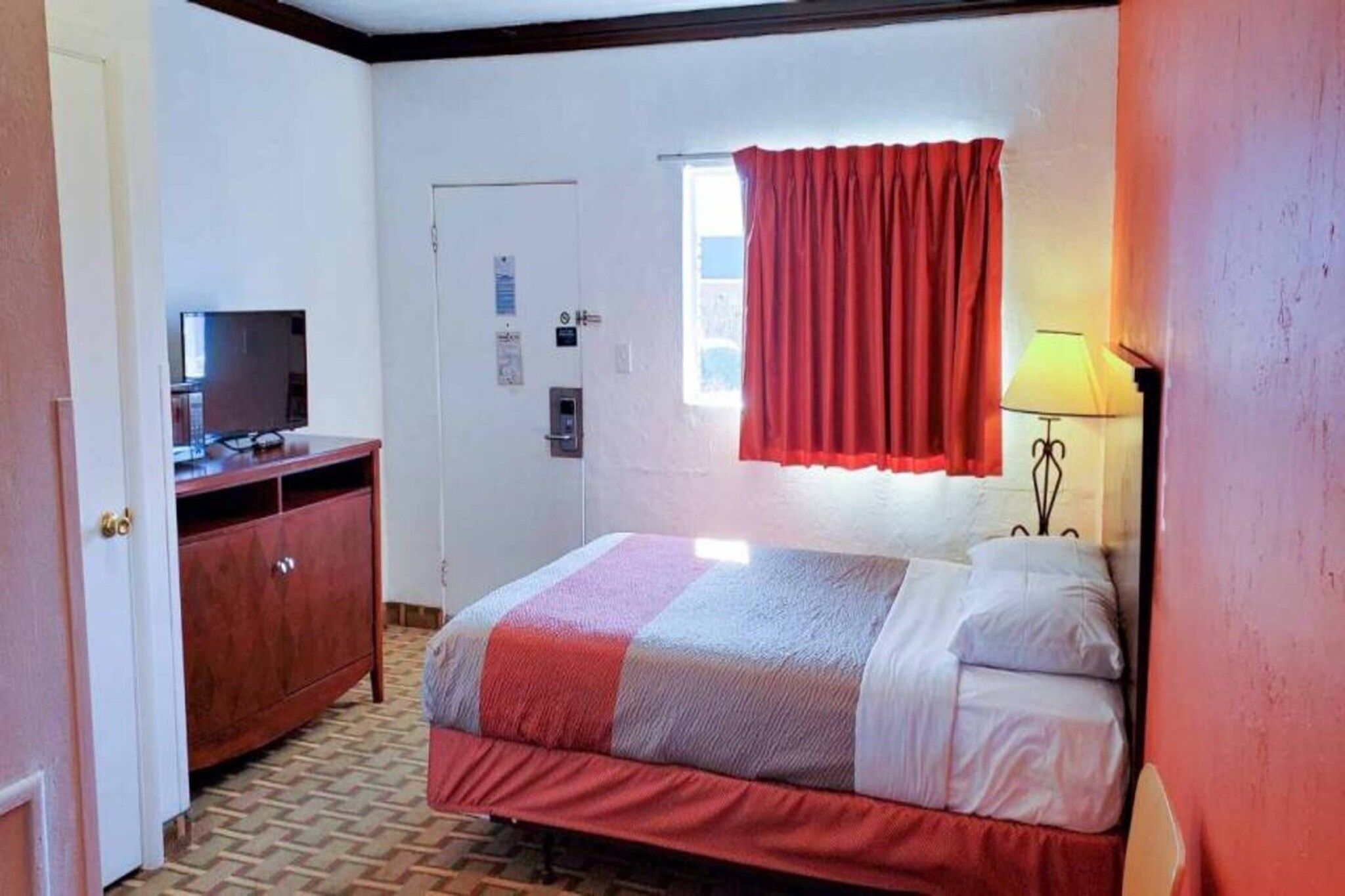 A hotel room with a bed, a desk, a television, a window with curtains, and a lamp.