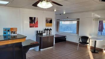 A hotel room with a kitchenette, a dining area, a ceiling fan, and a window with blinds.