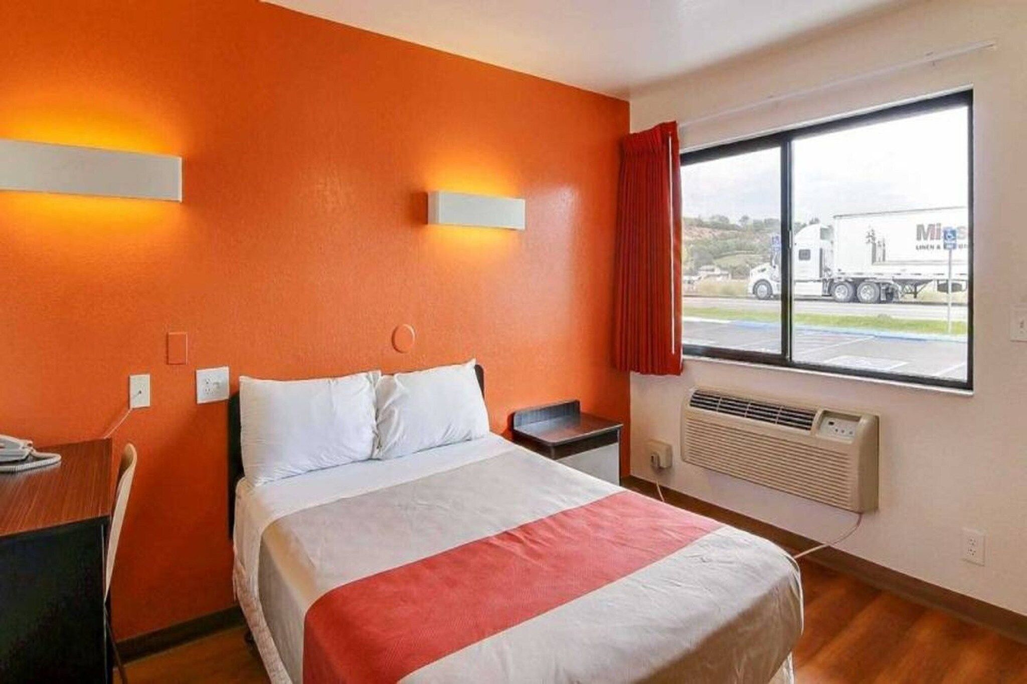 A hotel room with a bed, a desk, a chair, a window with a view of a truck, and a wall-mounted air conditioner.