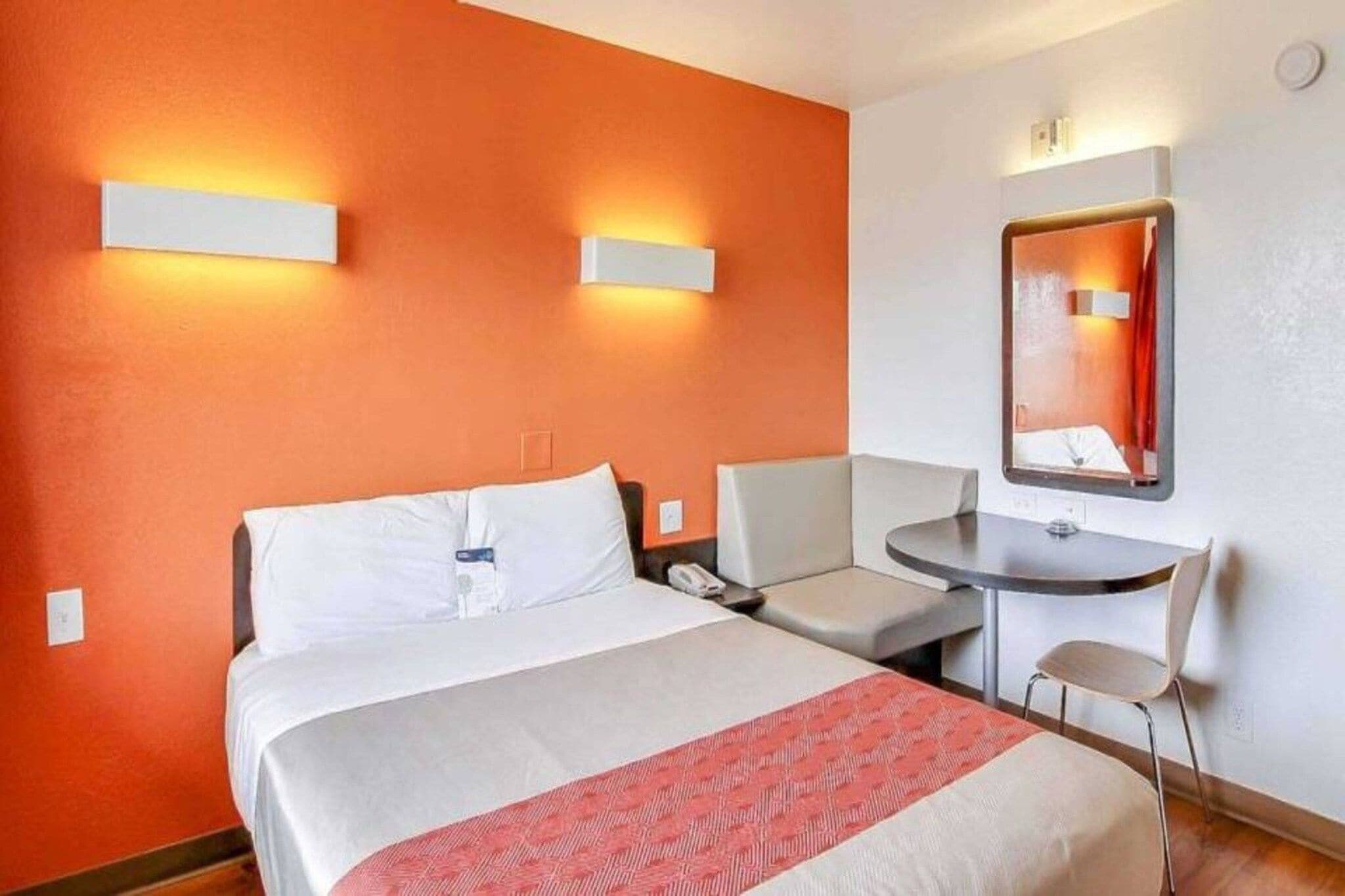 A hotel room with a bed, bedside table, chair, and a small desk with a mirror.