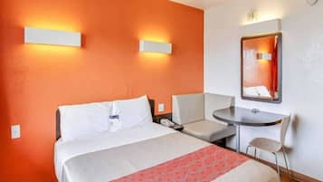 A hotel room with a bed, bedside table, chair, and a small desk with a mirror.
