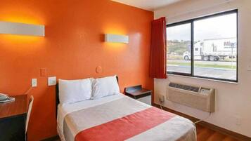 A hotel room with a bed, a desk, a chair, a window with a view of a truck, and a wall-mounted heater.