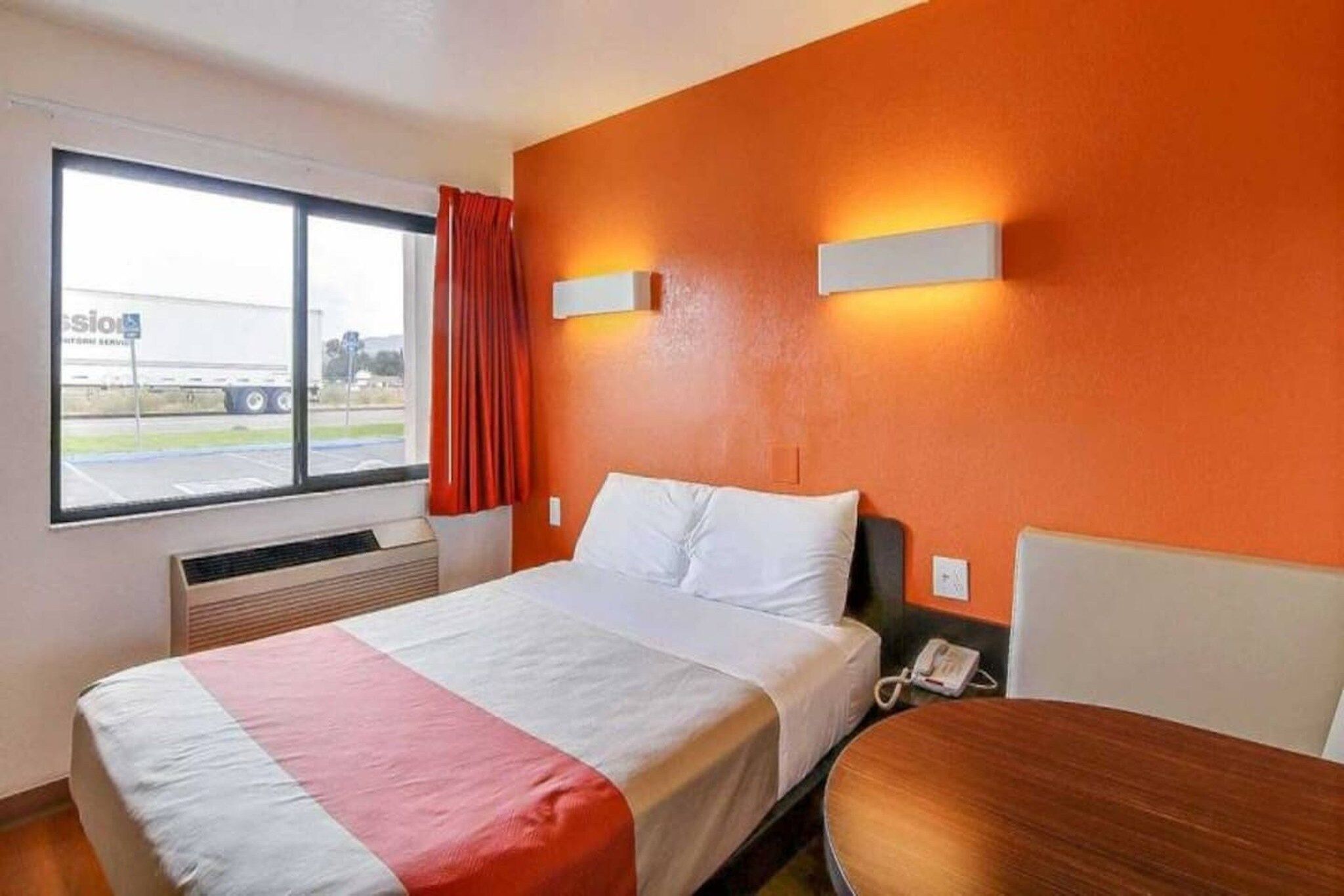 Habitación de hotel con cama, mesita de noche, ventana con vista a un camión y teléfono instalado en la pared.