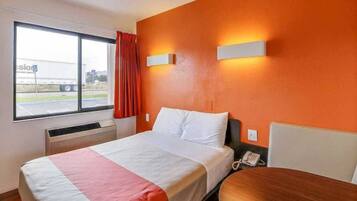 Habitación de hotel con cama, mesita de noche, ventana con vista a un camión y teléfono instalado en la pared.