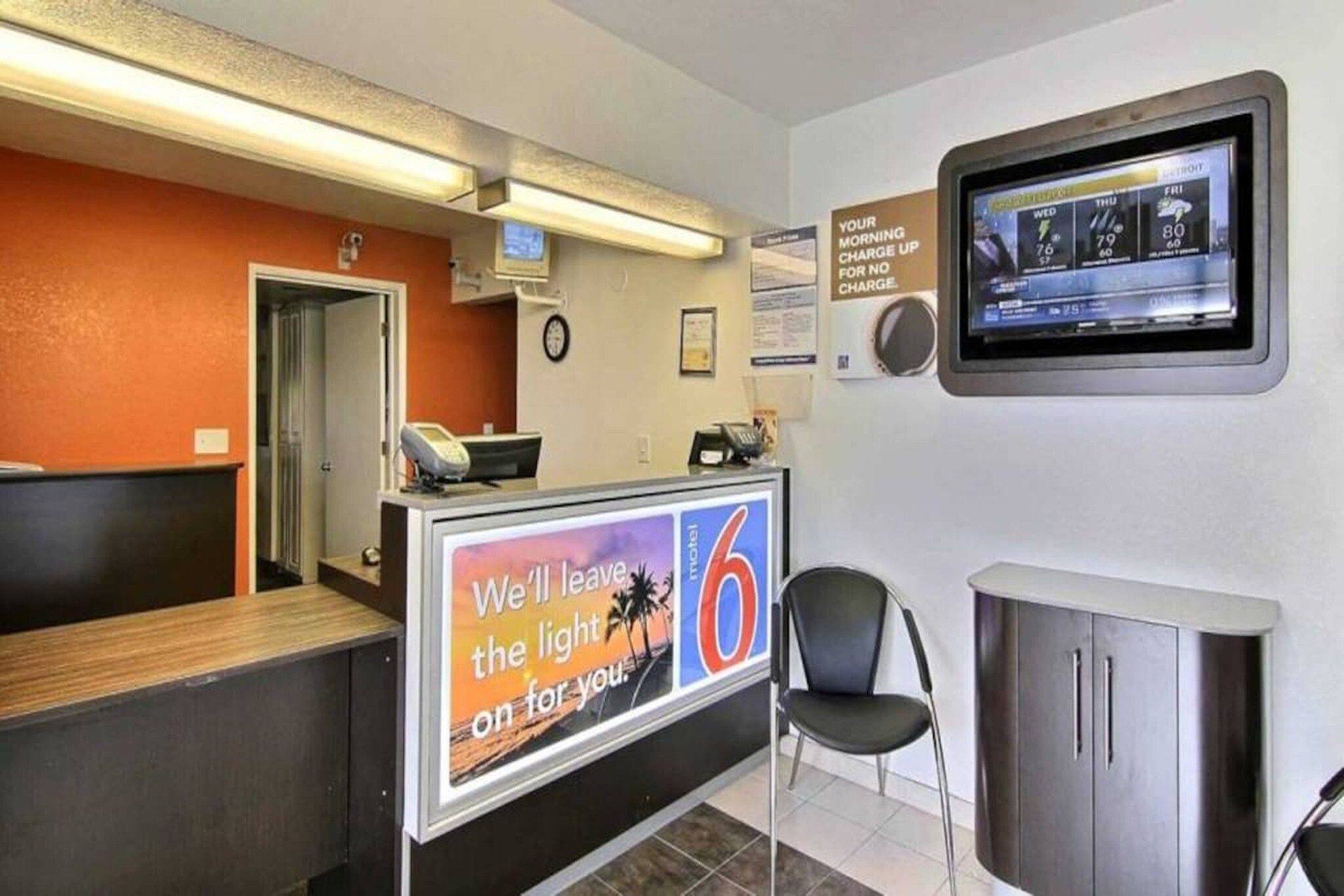 Recepção de hotel com painel digital, balcão, cadeira e armário.