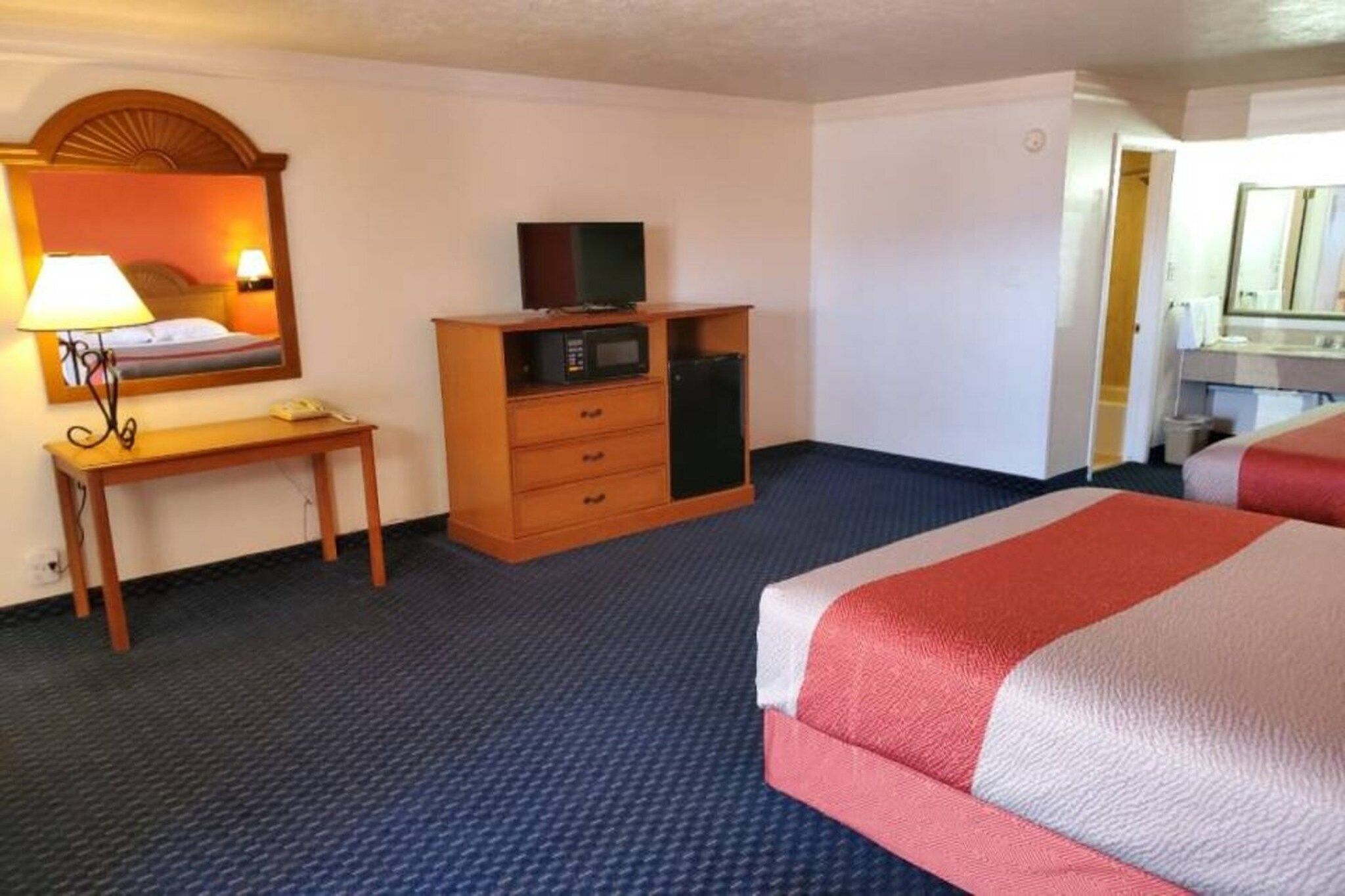 Habitación de hotel con cama, escritorio con lámpara, cómoda con televisor y microondas, y baño visible a través de una puerta abierta.