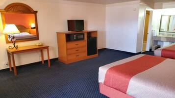 Habitación de hotel con cama, escritorio con lámpara, cómoda con televisor y microondas, y baño visible a través de una puerta abierta.