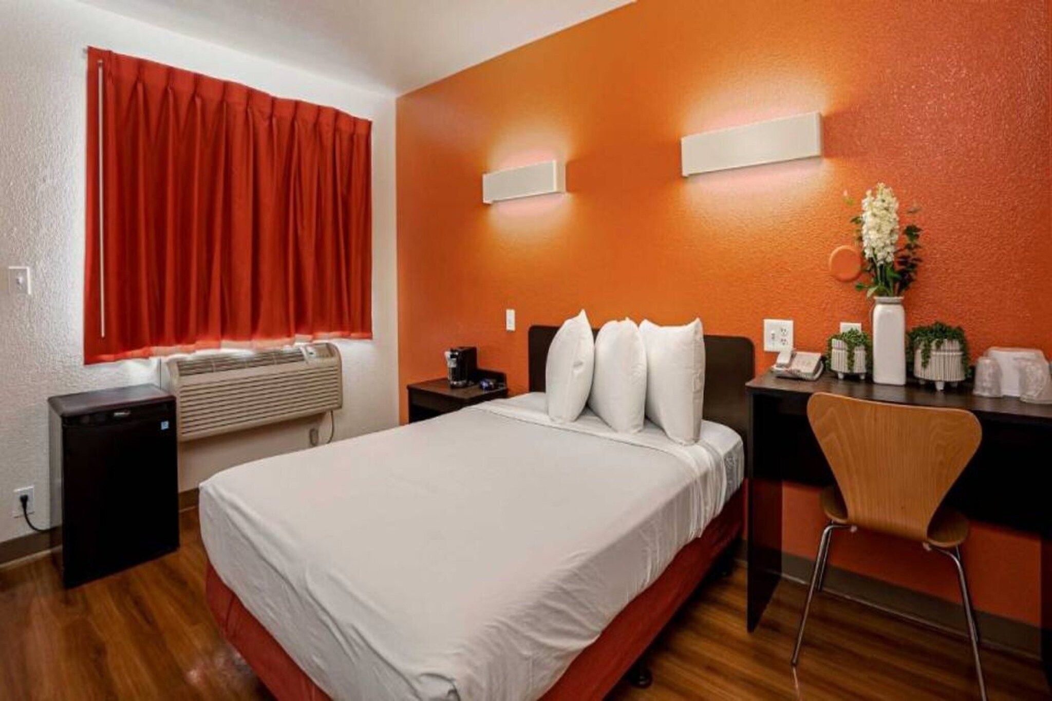 A hotel room with a bed, a desk, a chair, a desk lamp, a telephone, and a vase with flowers.