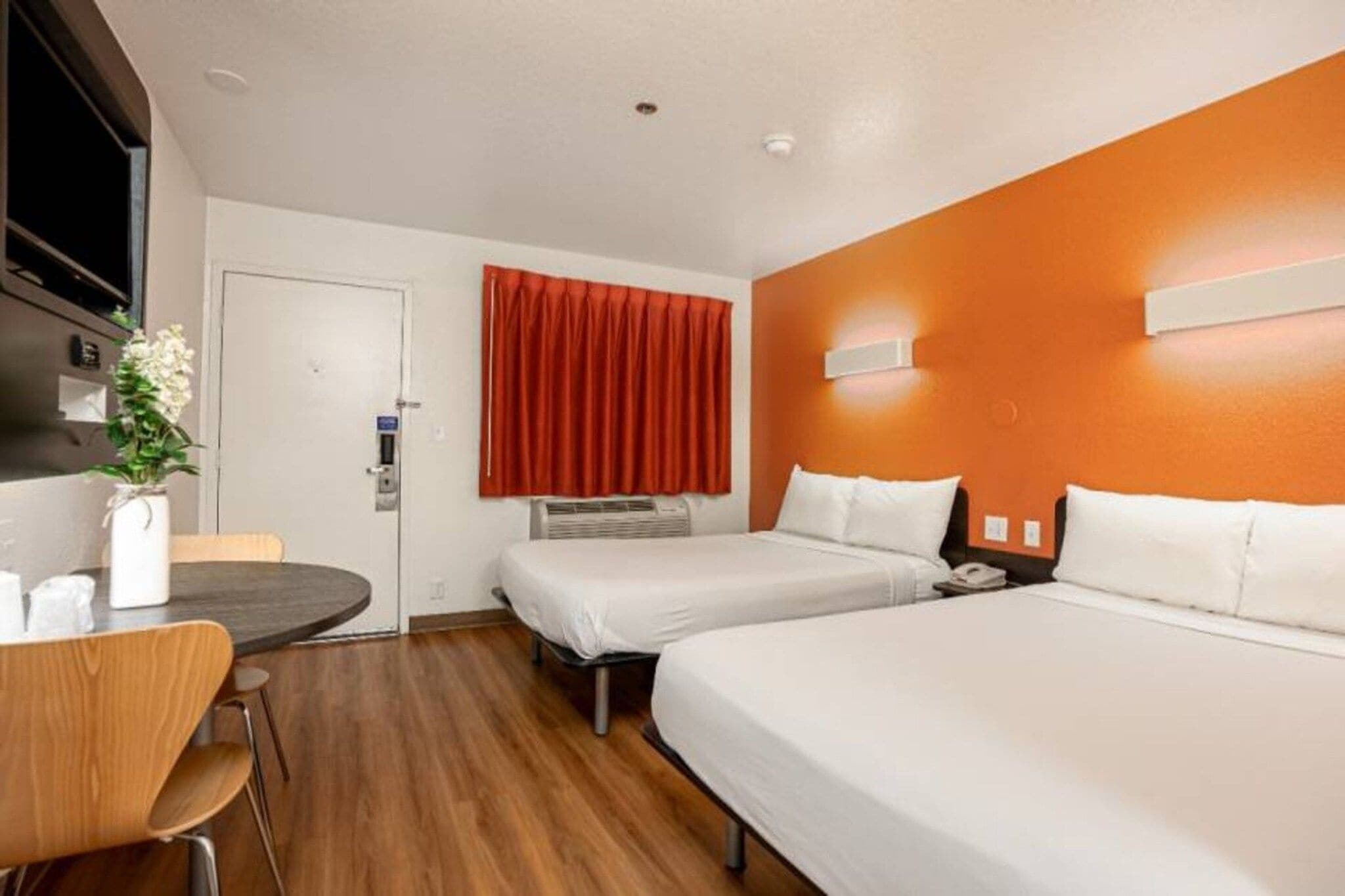 A hotel room with two beds, a desk, a chair, a vase with flowers, and a red curtain.