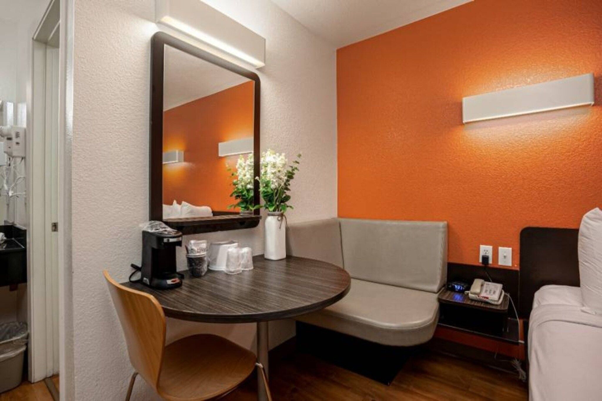 A hotel room with a round table, a chair, a sofa, a bedside table with a phone, and a mirror.
