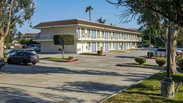 A motel with a central parking lot, a row of single-story buildings with balconies, and a small tree in the foreground.