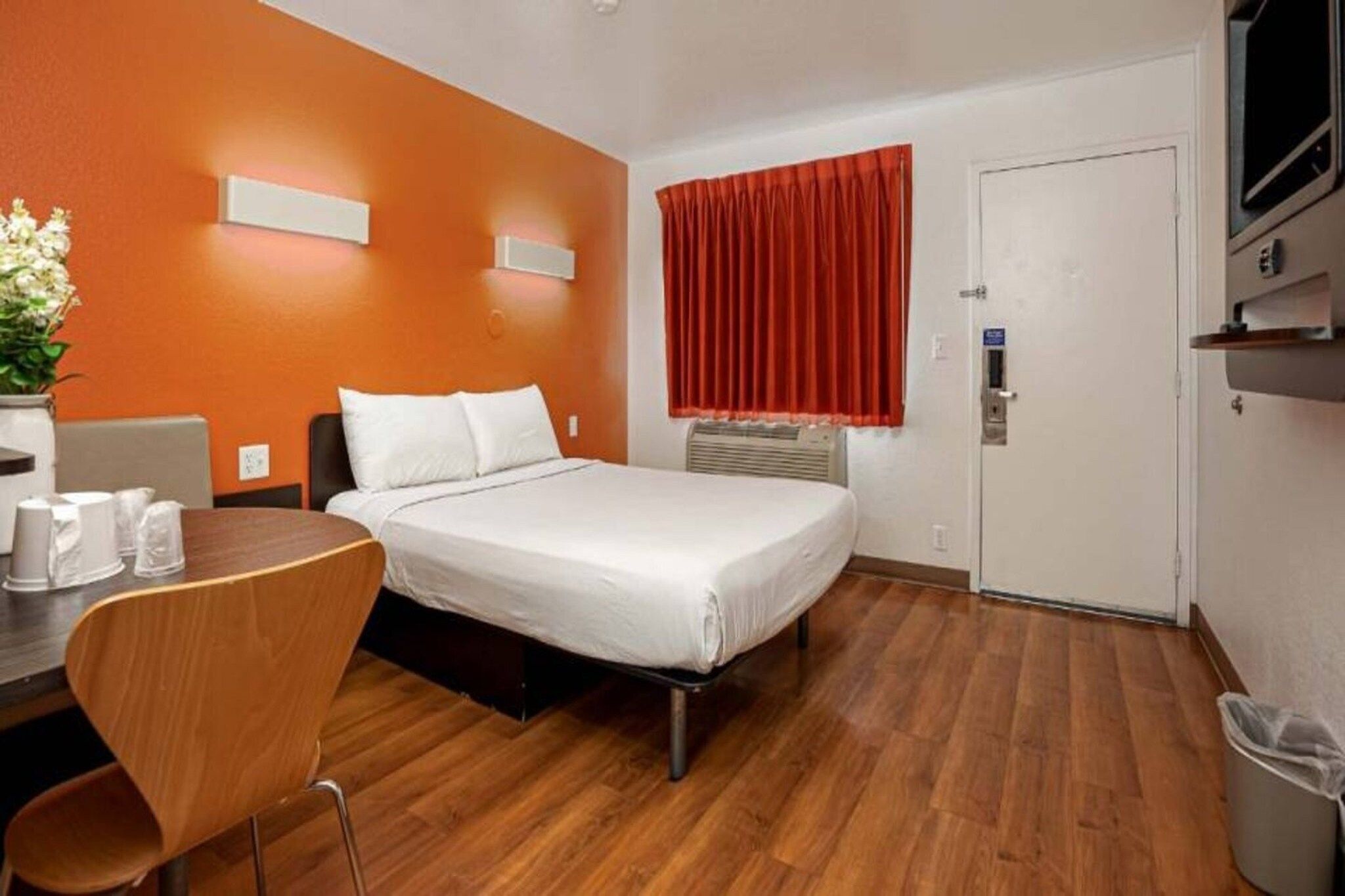 A hotel room with a bed, a desk, a chair, a trash bin, and a door.