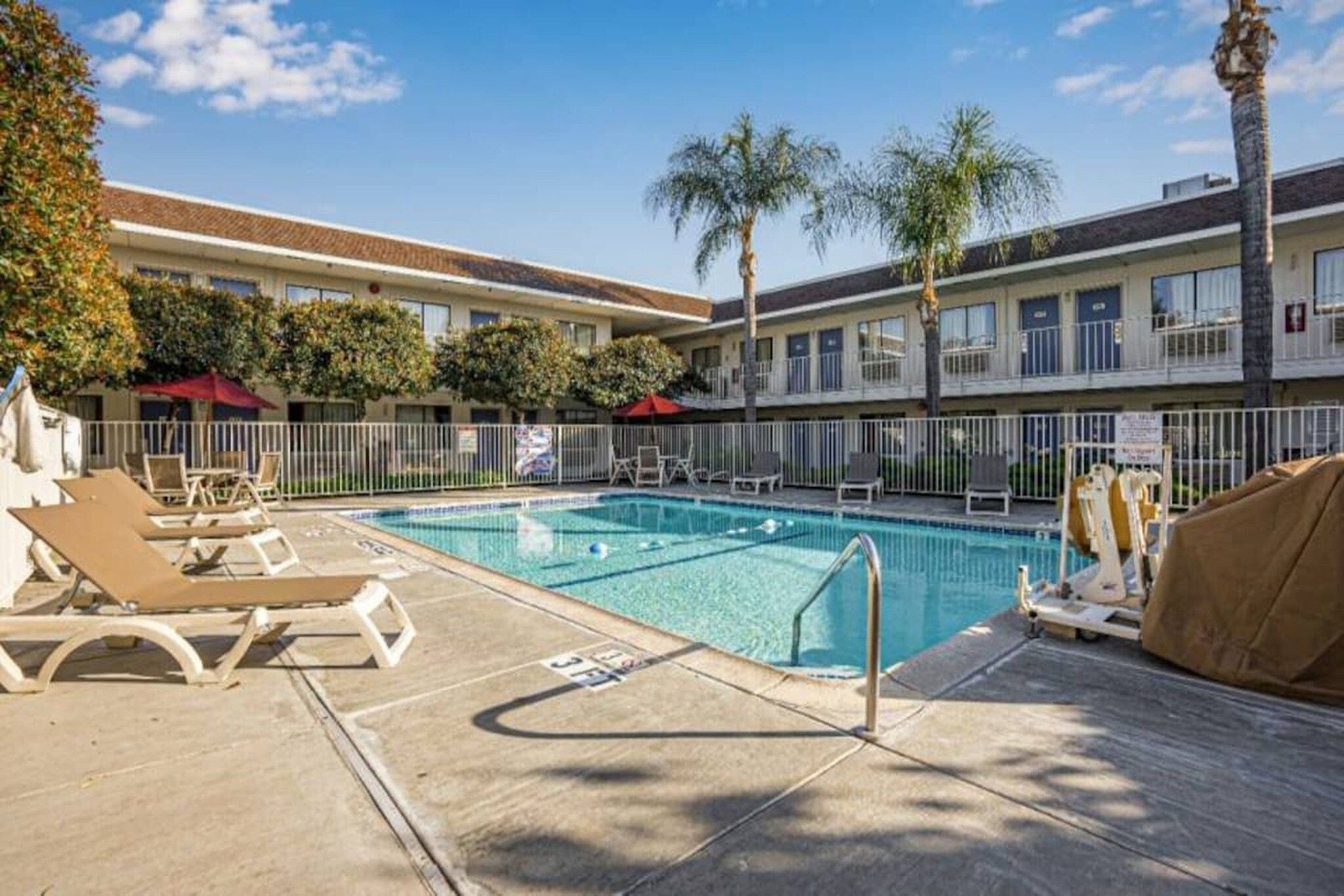 Une piscine avec des chaises longues, un espace délimité par une clôture et, au fond, un bâtiment doté de nombreuses fenêtres.
