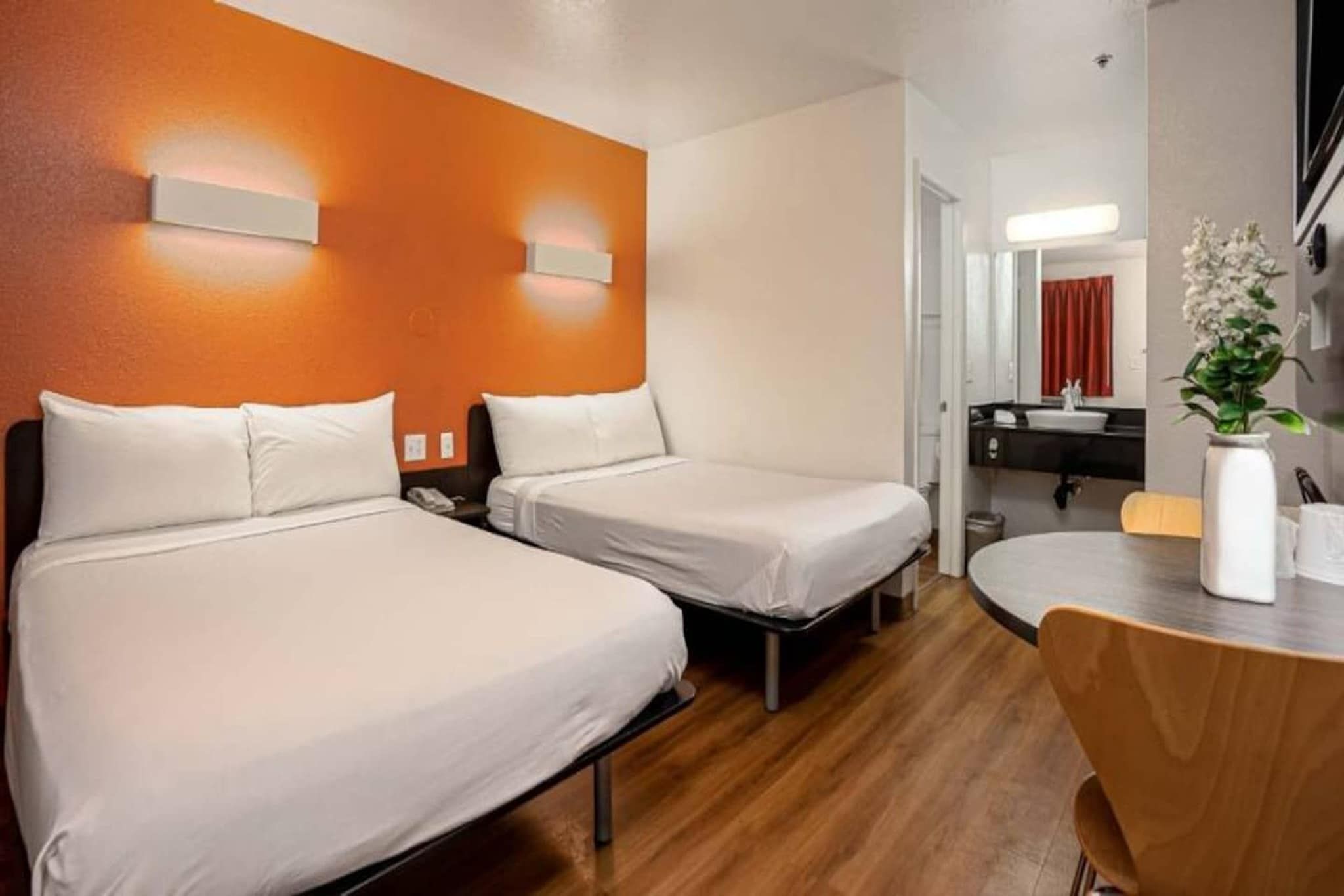 A hotel room with two beds, a wooden floor, a round table with chairs, and a bathroom visible through an open door.