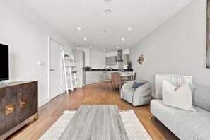 Een moderne woonkamer met een bank, een salontafel en een televisie. Achtergrond is een keuken met kastjes en een aanrecht.