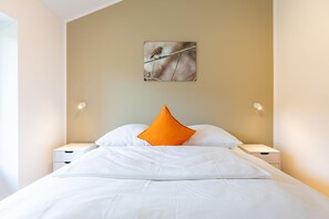 Ein Hotelzimmer mit einem Bett, weißer Bettwäsche, einem orangefarbenen Kissen, zwei Nachttischen und einem Wandbild.