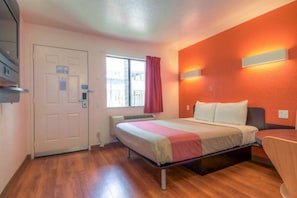 Ein Hotelzimmer mit Bett, Schreibtisch, einem fenster mit Vorhängen, einer Tür und einer Wandleuchte.
