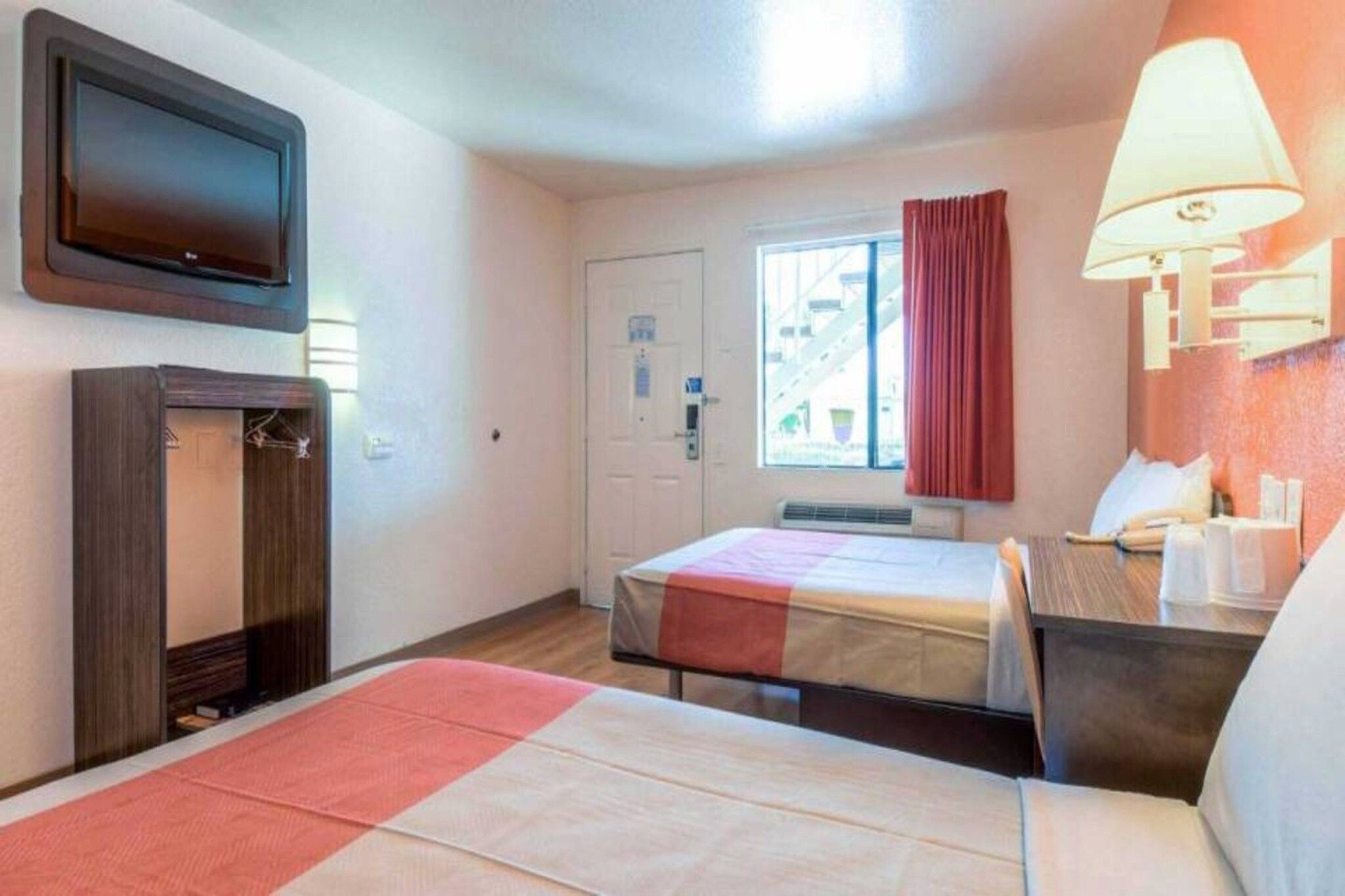 A hotel room with a bed, a desk, a television, a window with curtains, and a wall-mounted lamp.