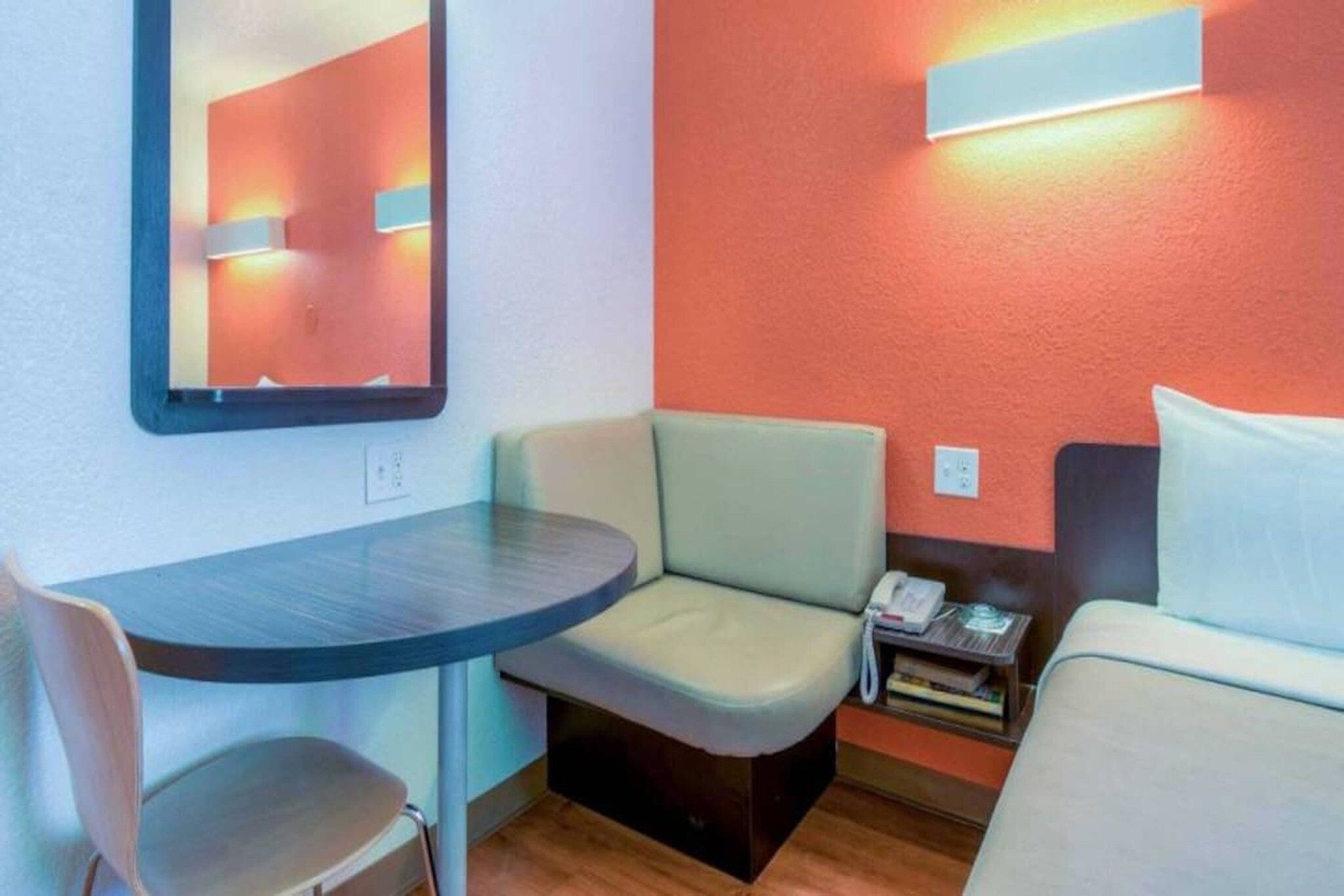 Ein kleines Hotelzimmer mit einem Einzelbett, einem Tisch, einem Stuhl, einem Nachttisch mit Telefon und einem an der Wand befestigten Spiegel.