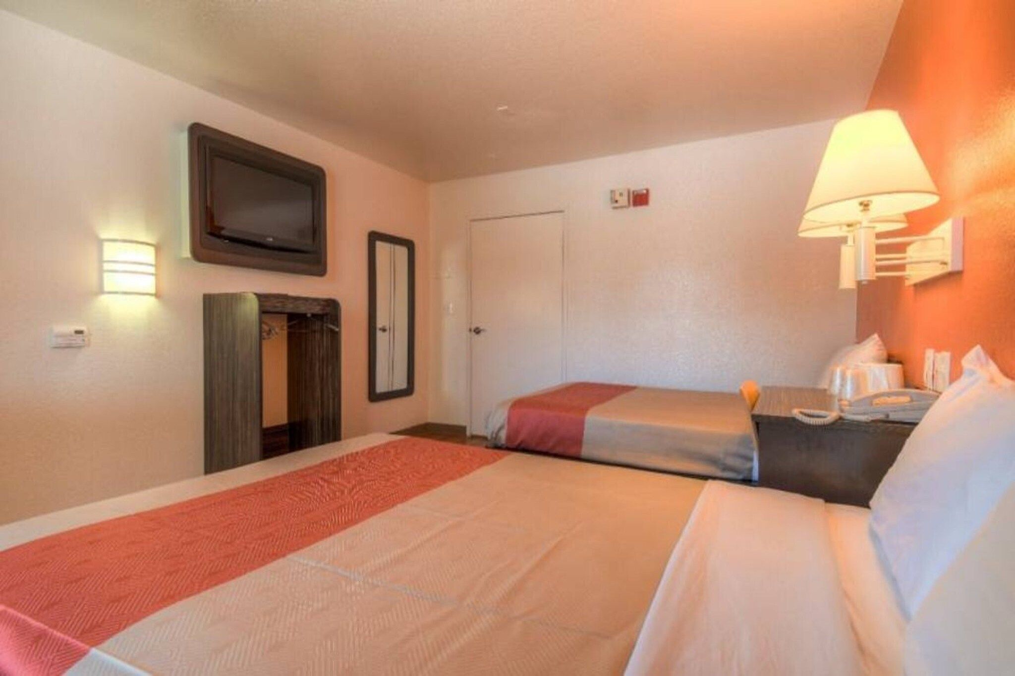 A hotel room with two beds, a television, a door, and a wall-mounted light fixture.