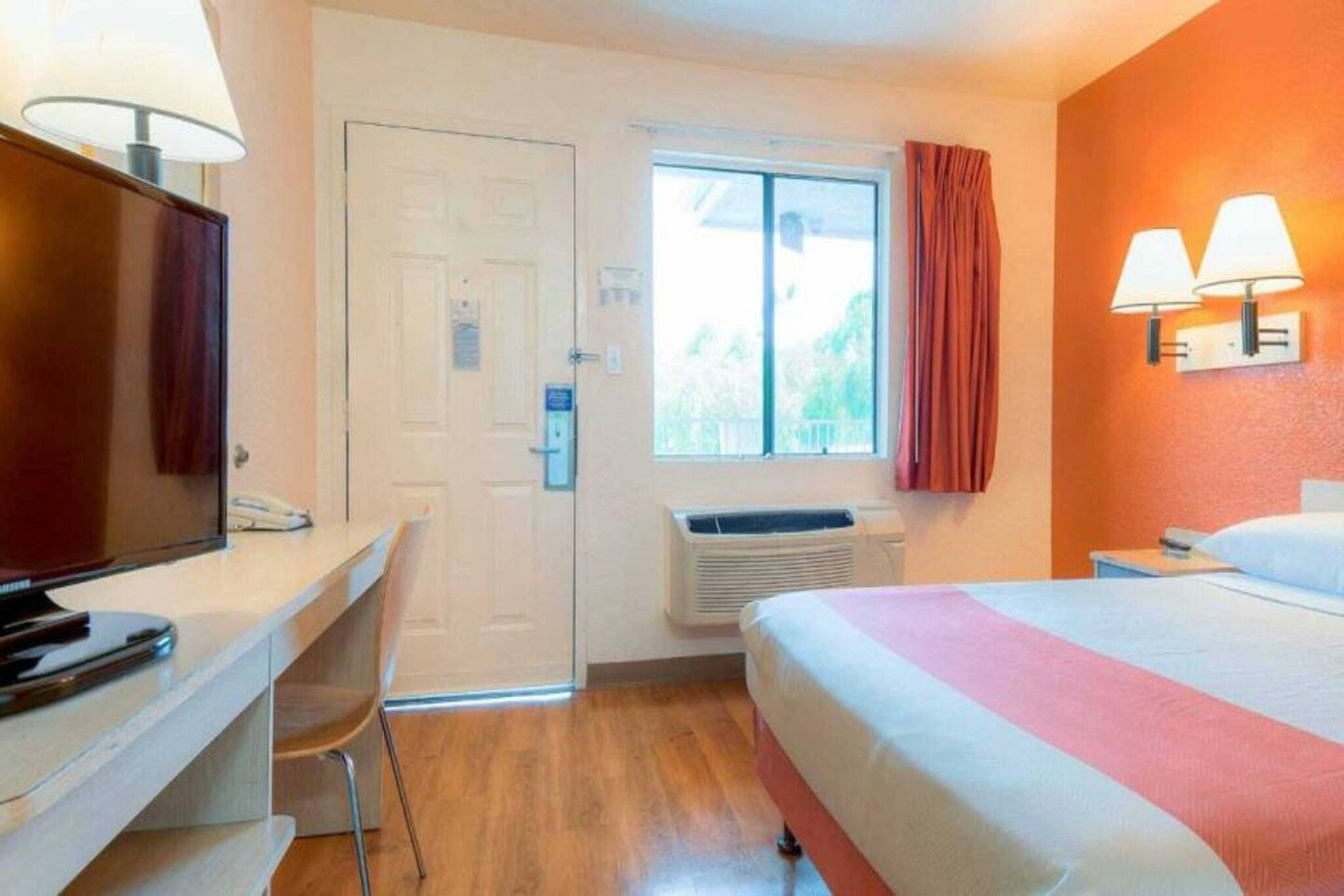 Habitación de hotel con cama, escritorio, televisor, ventana con cortinas y aire acondicionado.