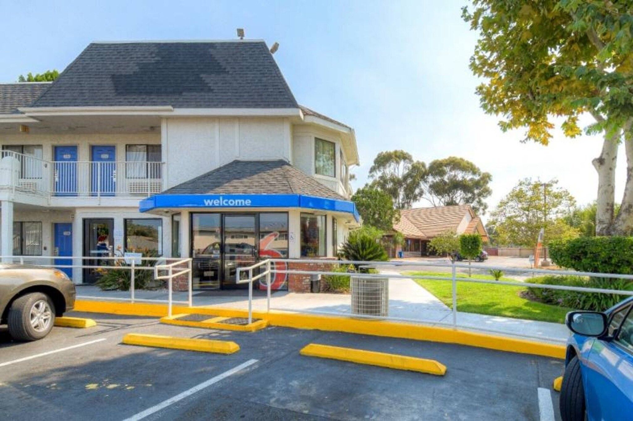 A hotel entrance with a 'welcome' sign, a covered waiting area, and a parking lot with yellow curbs.