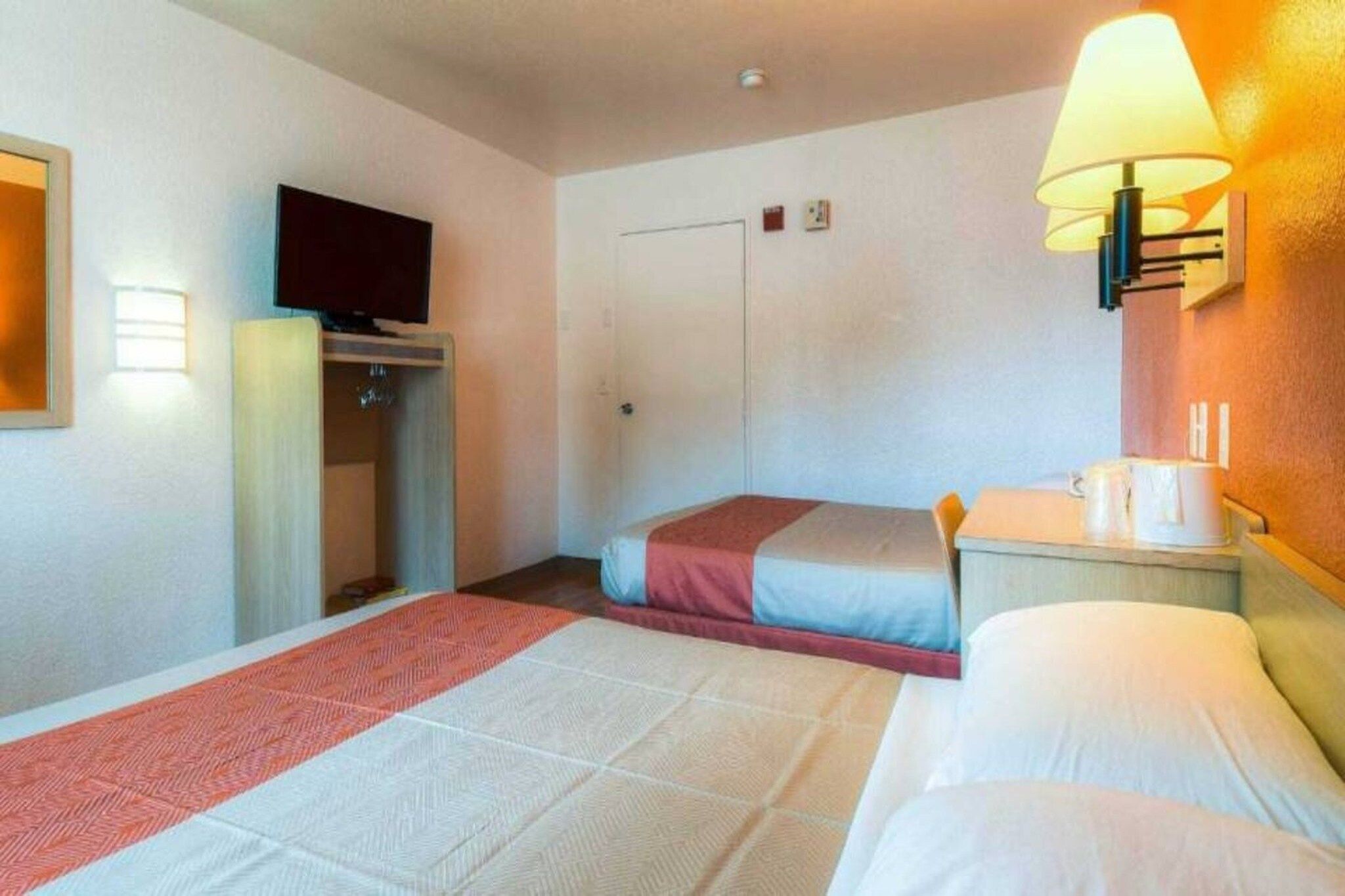 A hotel room with two beds, a television, a bedside table with a lamp, and a small desk with a chair.
