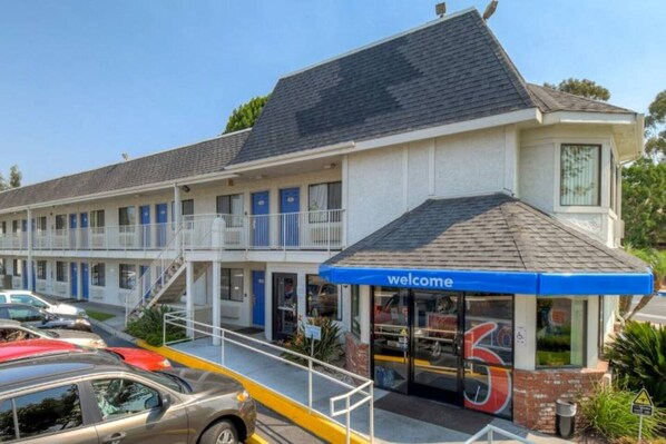 Un motel avec une enseigne « Bienvenue », une entrée abritée et un parking où sont garées des voitures.