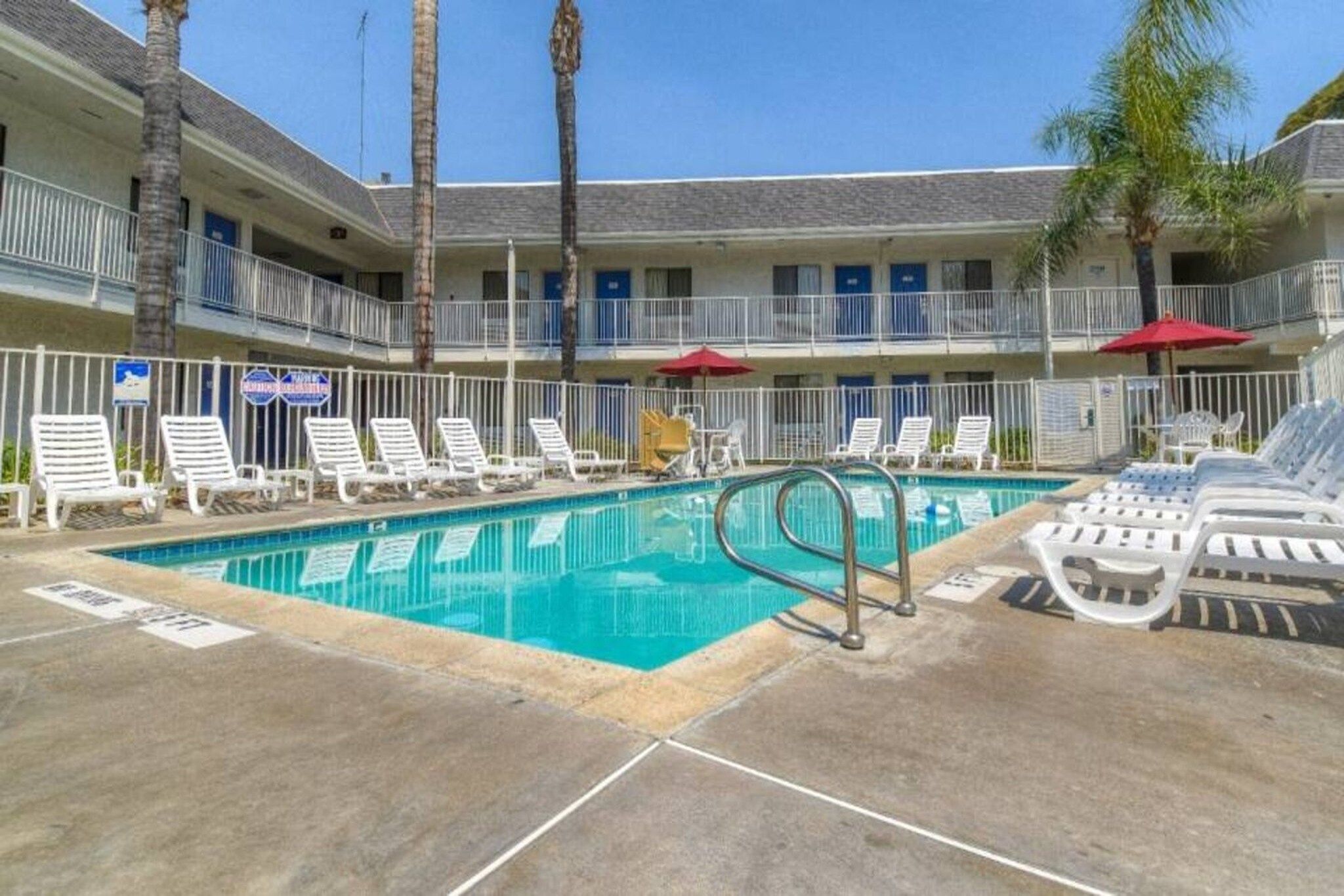 A hotel pool area with multiple lounge chairs and umbrellas, surrounded by a white fence.