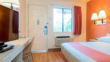 A hotel room with a bed, a desk, a television, a window with curtains, and an air conditioning unit.