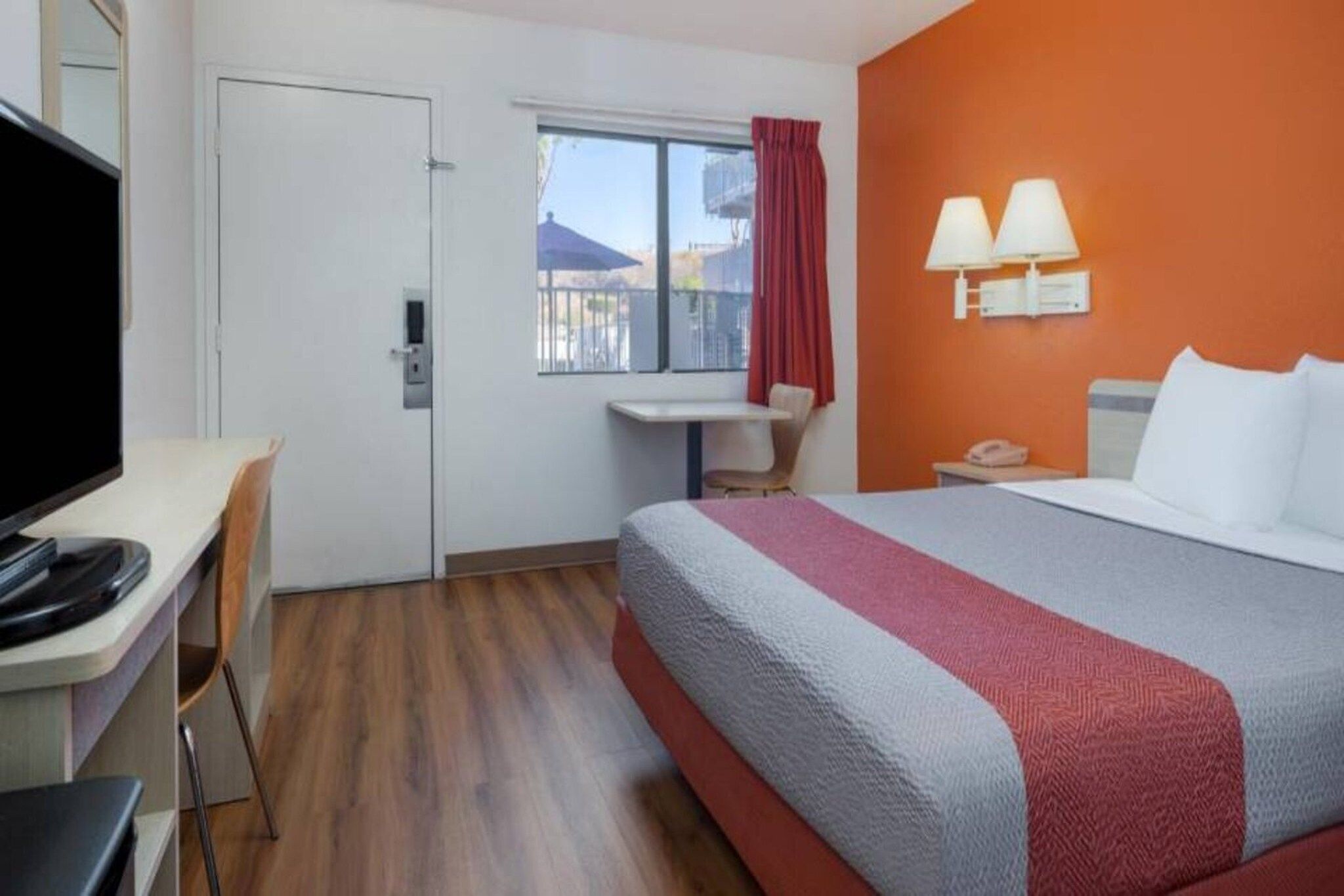 Una habitación de hotel con cama, escritorio, silla, televisión, ventana con cortinas y mesita de noche con lámpara.