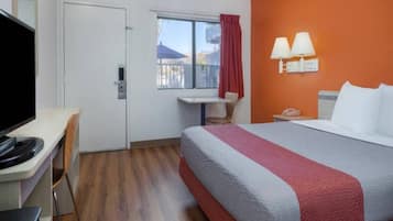 Una habitación de hotel con cama, escritorio, silla, televisión, ventana con cortinas y mesita de noche con lámpara.