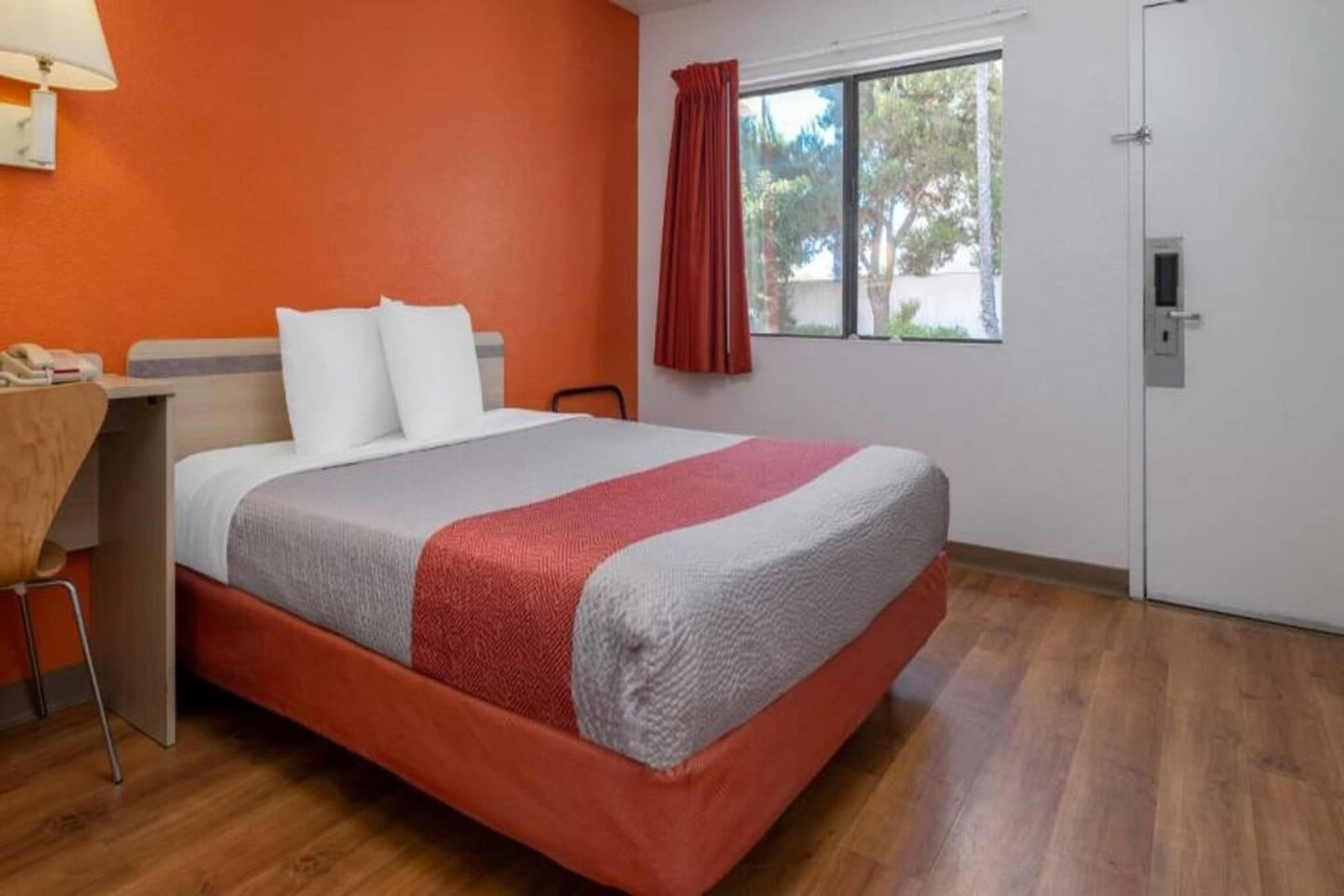 A hotel room with a bed, a desk, a chair, a window with curtains, and a door.