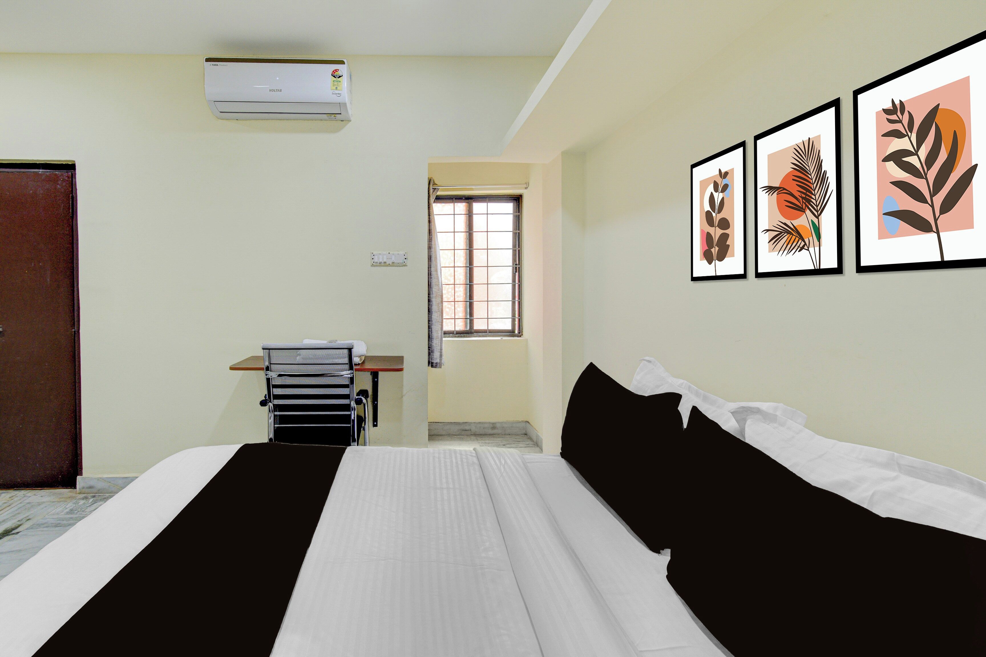 A hotel room with a bed, a desk, an air conditioner, a window, and framed artwork on the wall.