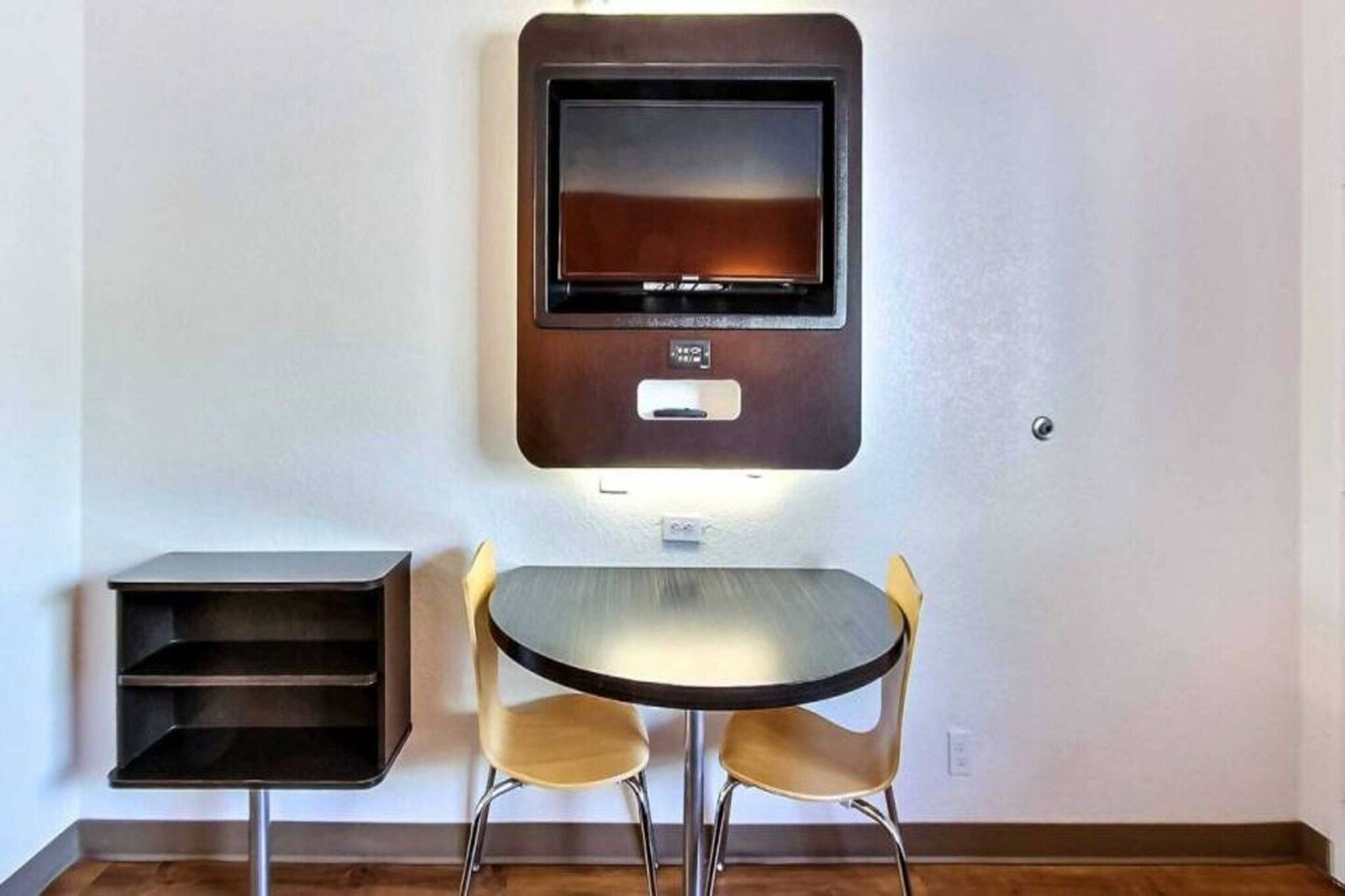 A room with a wall-mounted TV, a small table with two chairs, and a shelf unit.