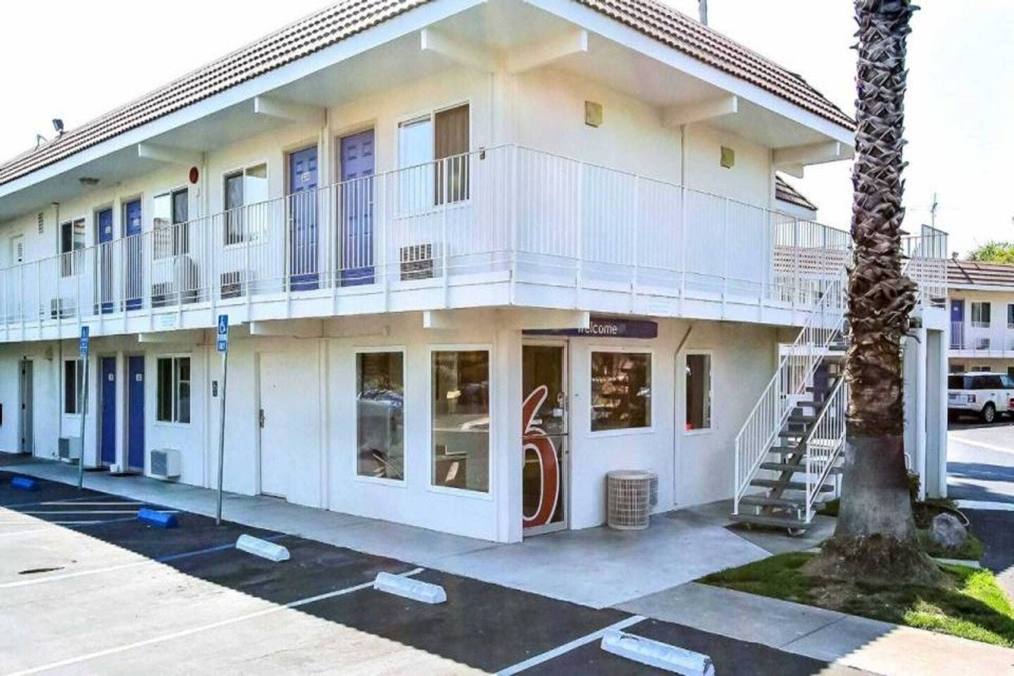 A two-story motel with a balcony on the upper floor, a visible entrance, and a palm tree in the foreground.