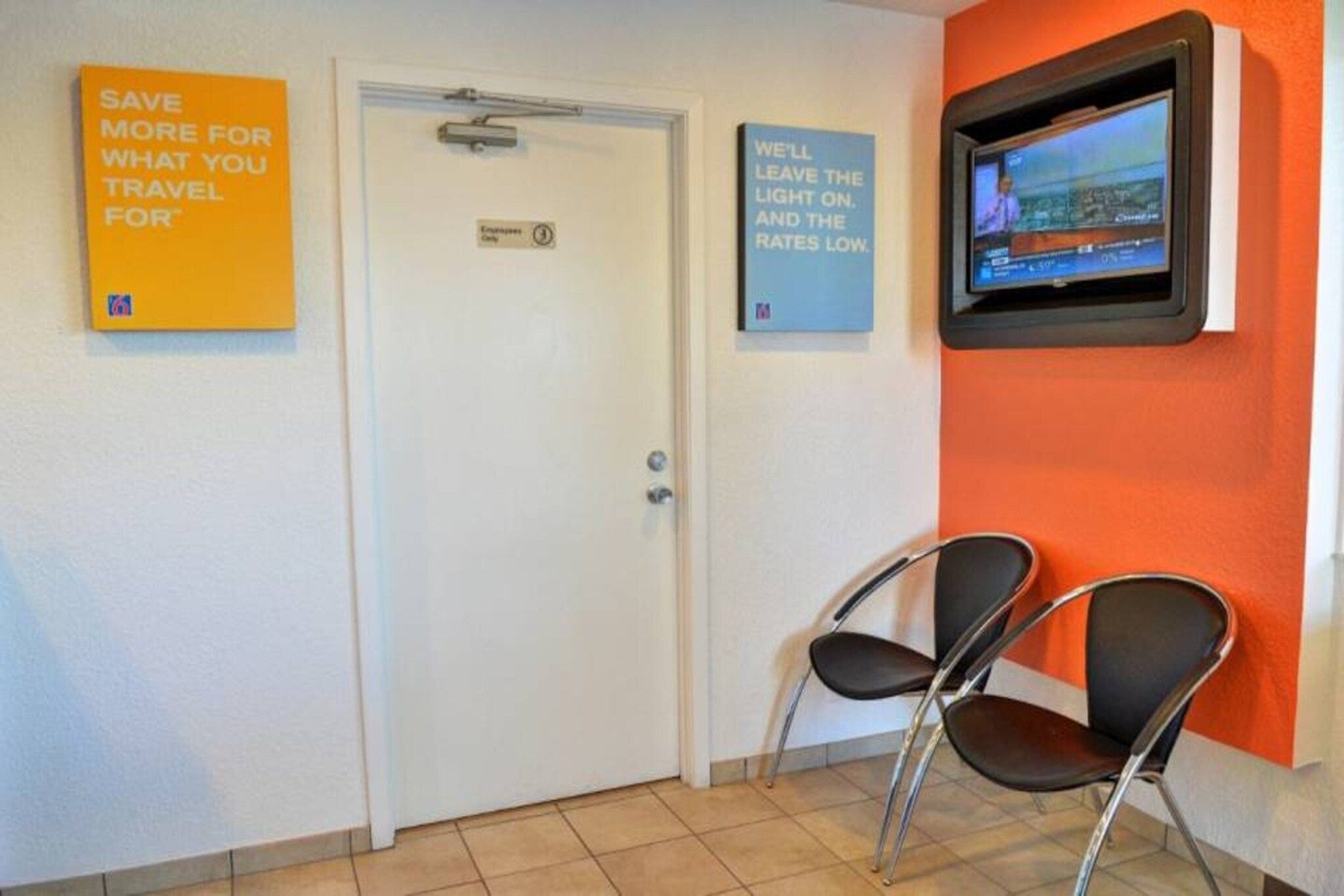 A hotel reception area with a closed door, two chairs, and a television mounted on the wall displaying a news channel.