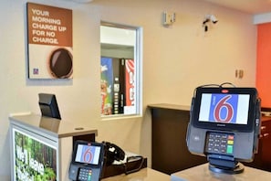 A hotel reception area with a digital kiosk displaying the number 6, a telephone, and a sign that reads 'YOUR MORNING CHARGE UP FOR NO CHARGE.'