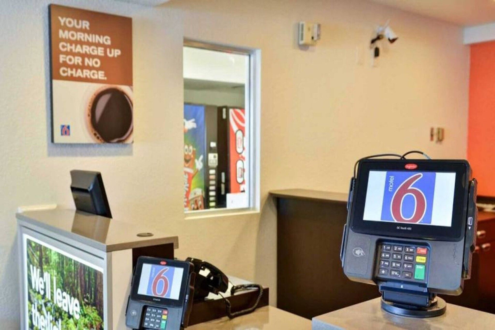 Een hotel receptie met een digitale kiosk waarop het cijfer 6 staat, een telefoon en een bord met de tekst 'OPLAADBEURT ZONDER KOSTEN'.