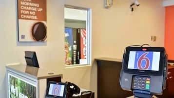 Een hotel receptie met een digitale kiosk waarop het cijfer 6 staat, een telefoon en een bord met de tekst 'OPLAADBEURT ZONDER KOSTEN'.